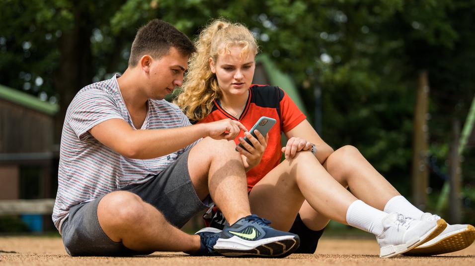 Zwei Jugendliche sitzen auf dem Boden und betrachten gemeinsam ein Smartphone. Ein Mädchen trägt ein rotes sportliches Oberteil.